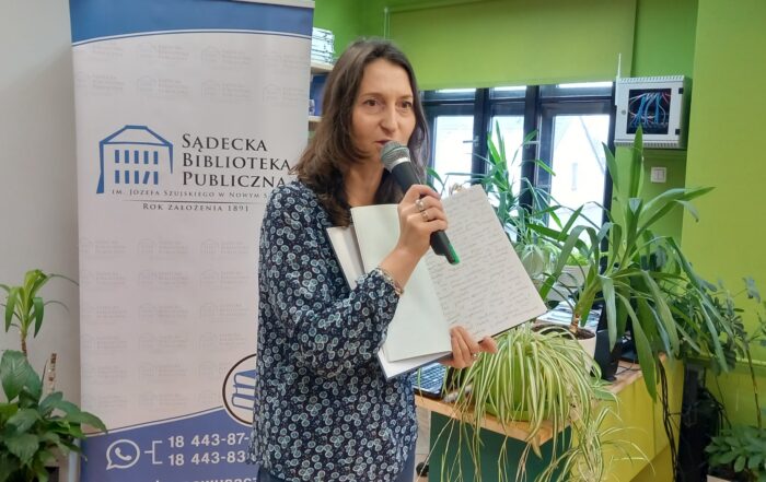 W pomieszczeniu, blisko nas, stoi kobieta z mikrofonem w prawej ręce. W lewej trzyma otwarty, duży i zapisany drobnym drukiem zeszyt. Wokół niej dużo roślin. W tle duży napis Sądecka Biblioteka Publiczna.