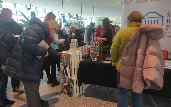 W wielkim pomieszczeniu grupa ludzi. Po lewej stronie kobieta pokazana od tyłu. Przez prawe ramię ma przewieszoną kurtkę. Obok mężczyzna ukazany również od tyłu. Sięga do białek skrzynki pełnej książek. W prawej ręce trzyma książkę.