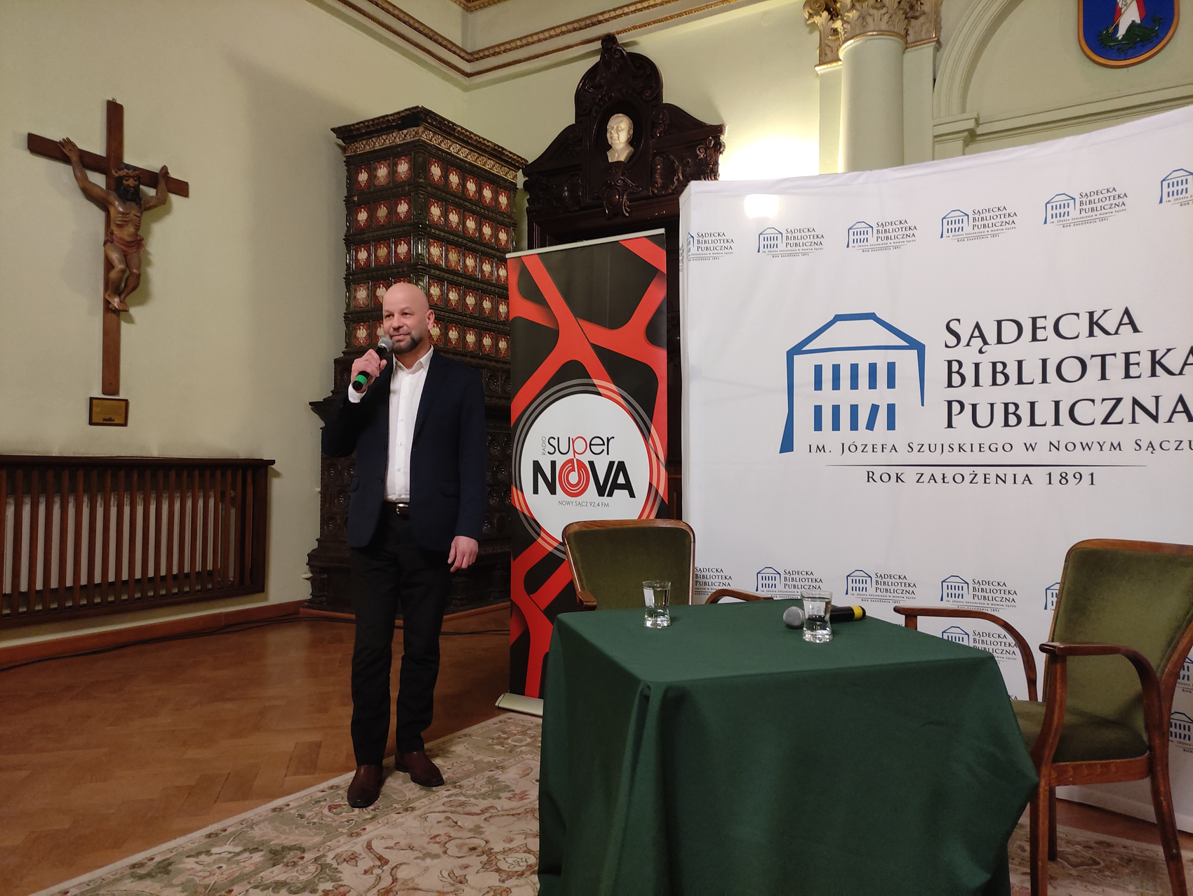 Artur Chamski (1) Sala Ratuszowa. Po lewej stronie stoi mężczyzna. W prawej ręce trzyma mikrofon. Obok niego stoi stół z zielonym obrusem. Na stole stoją dwie szklanki z wodą i mikrofon. Za stołem znajdują się dwa krzesła. W tle ścianka Sądeckiej Biblioteki Publicznej oraz roll-up Radia Supernova.