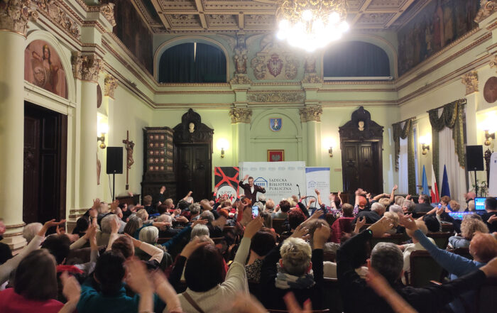 Grupa siedzących ludzi znajduje się w Sali Ratuszowej. Machają rękami. W oddali piosenkarz Artur Chamski. W tle ścianka Sądeckiej Biblioteki Publicznej i dwa roll-upy: Sądeckiej Biblioteki Publicznej i Radia Supernova.