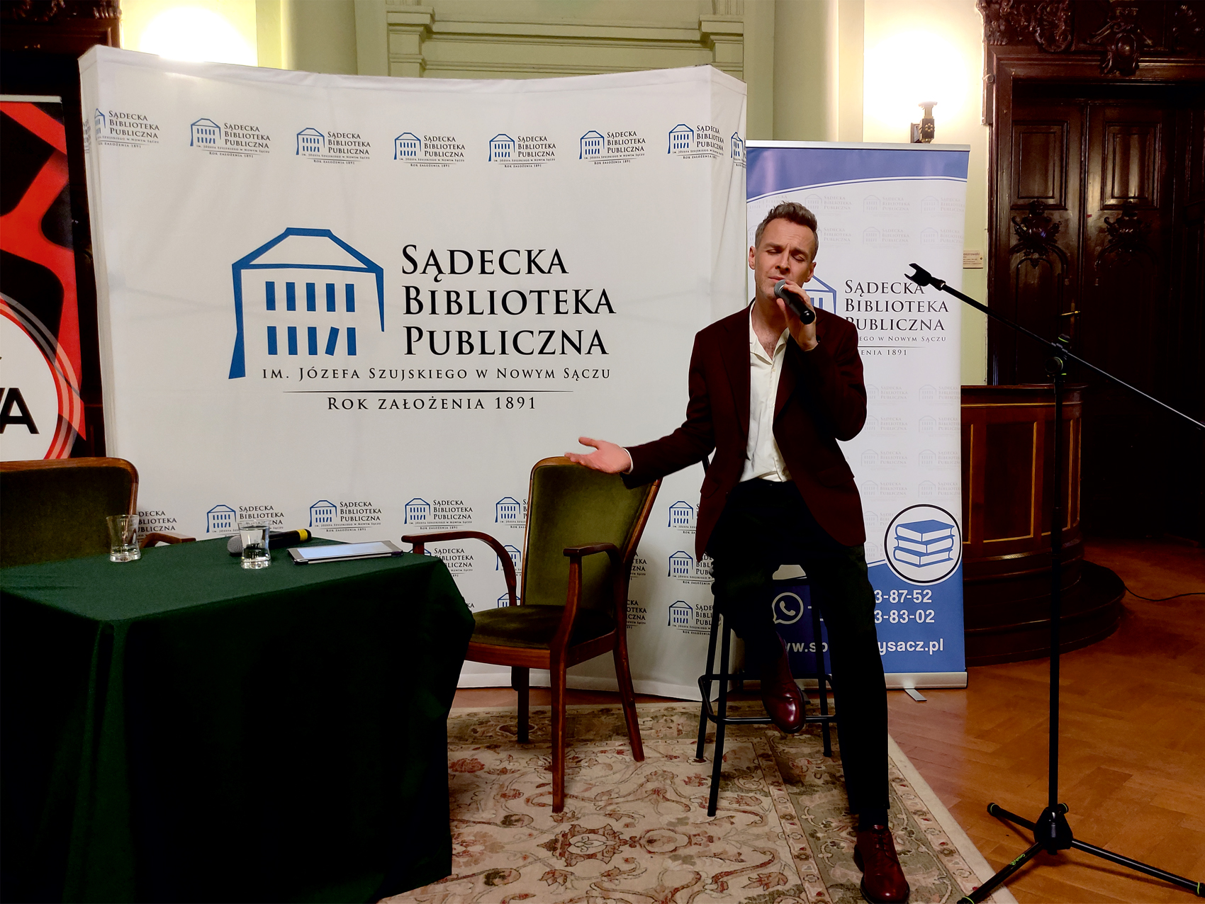 Artur Chamski (12) Po prawej stronie mężczyzna siedzi na krześle barowym. W prawej dłoni trzyma mikrofon i śpiewa do niego. Mężczyzna ma zamknięte oczy. Po lewej stronie stół z zielonym obrusem. Na stole dwie szklanki i mikrofon. W tle ścianka Sądeckiej Biblioteki Publicznej.