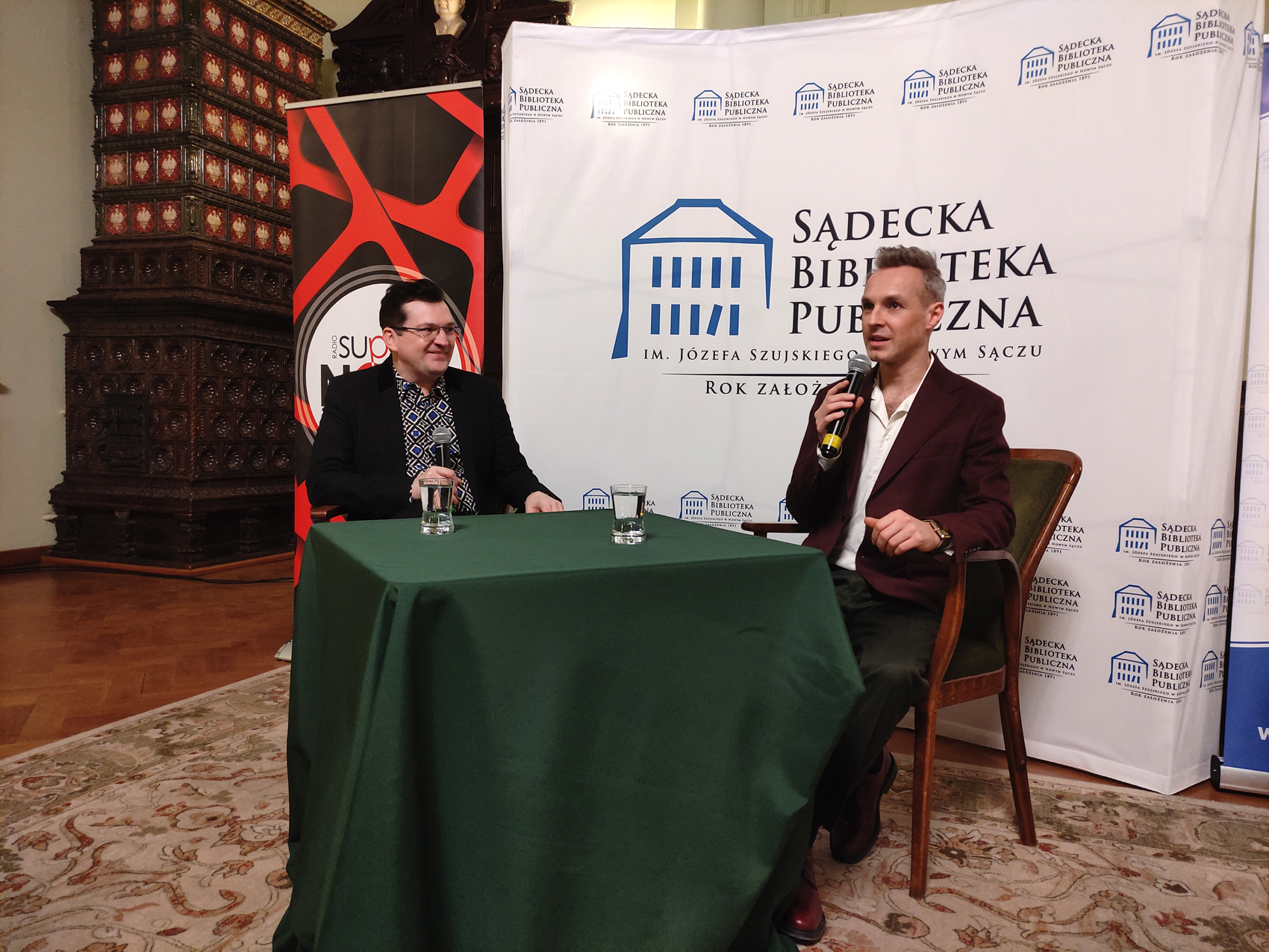 Artur Chamski (2) Sala Ratuszowa. Na środku stoi stół nakryty zielonym obrusem, a na nim dwie szklanki wody. Za stołem siedzą na krzesłach dwaj mężczyźni. Każdy z nich trzyma w prawej dłoni mikrofon. Mężczyzna po lewej stronie mówi do mikrofonu. Po lewej stronie stoi mężczyzna. W prawej ręce trzyma mikrofon. W tle ścianka Sądeckiej Biblioteki Publicznej oraz roll-up Radia Supernova.