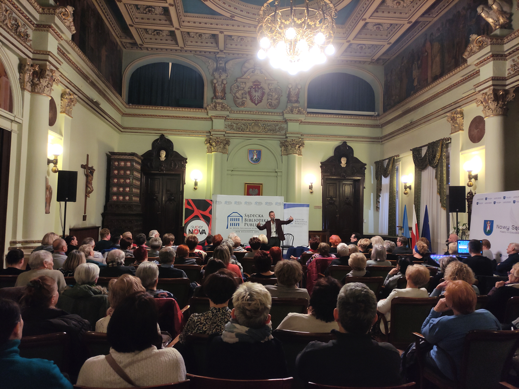 Artur Chamski (9) Sala Ratuszowa. Grupa siedzących ludzi, pokazana od tyłu. Przed nimi stoi mężczyzna, który śpiewa do mikrofonu na statywie. Obydwie ręce ma uniesione do góry. Za mężczyzną ścianka Sądeckiej Biblioteki Publicznej i dwa roll-upy: Sądeckiej Biblioteki Publicznej i Radia Supernova.