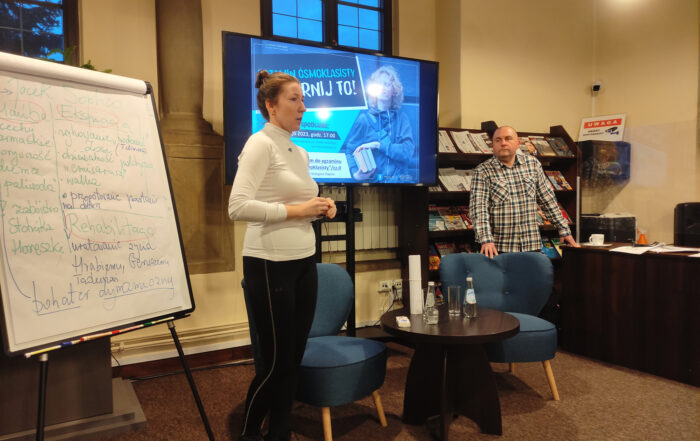 W pomieszczeniu stoją kobieta i mężczyzna. Obok kobiety, po lewej stronie stoi flipchart. Między kobietą a mężczyzną stoi stolik z dwoma niskimi fotelami. W duży ekran, regał z gazetami i biurko.