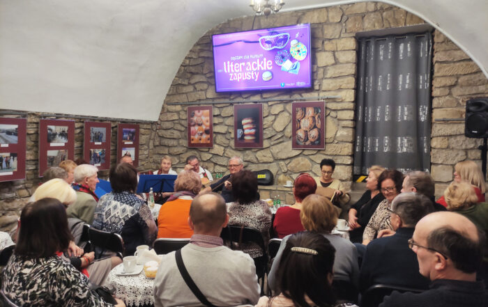 Grupa siedzących na krzesłach ludzi, zwróconych tyłem. Przed nimi siedzi na krzesłach grupa ludzi z zespołu muzycznego. Każdy z trzech siedzących mężczyzn trzyma instrument: akordeon, gitarę i skrzypce.