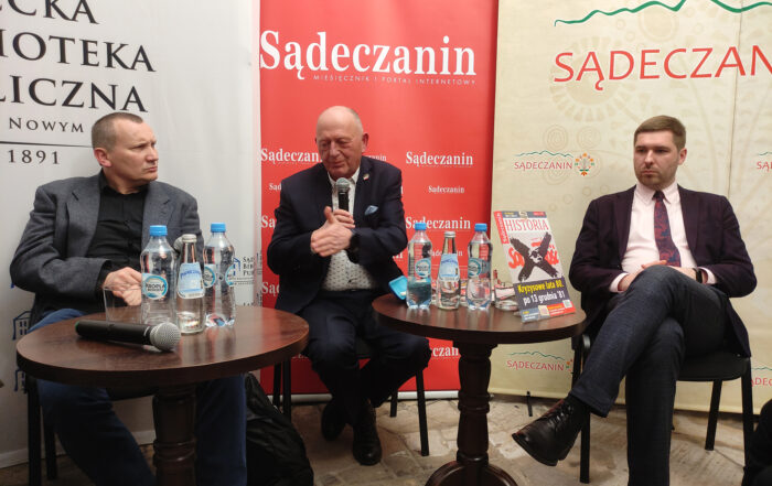 Na zdjęciu dwa okrągłe stoliki, a na nich butelki z wodą, szklanki, okładka czasopisma. Za stolikami siedzi na krzesłach trzech mężczyzn. Mężczyzna pośrodku trzyma w jednej ręce mikrofon, drugą ręką gestykuluje. W tle ścianki z napisem "Sądeczanin".