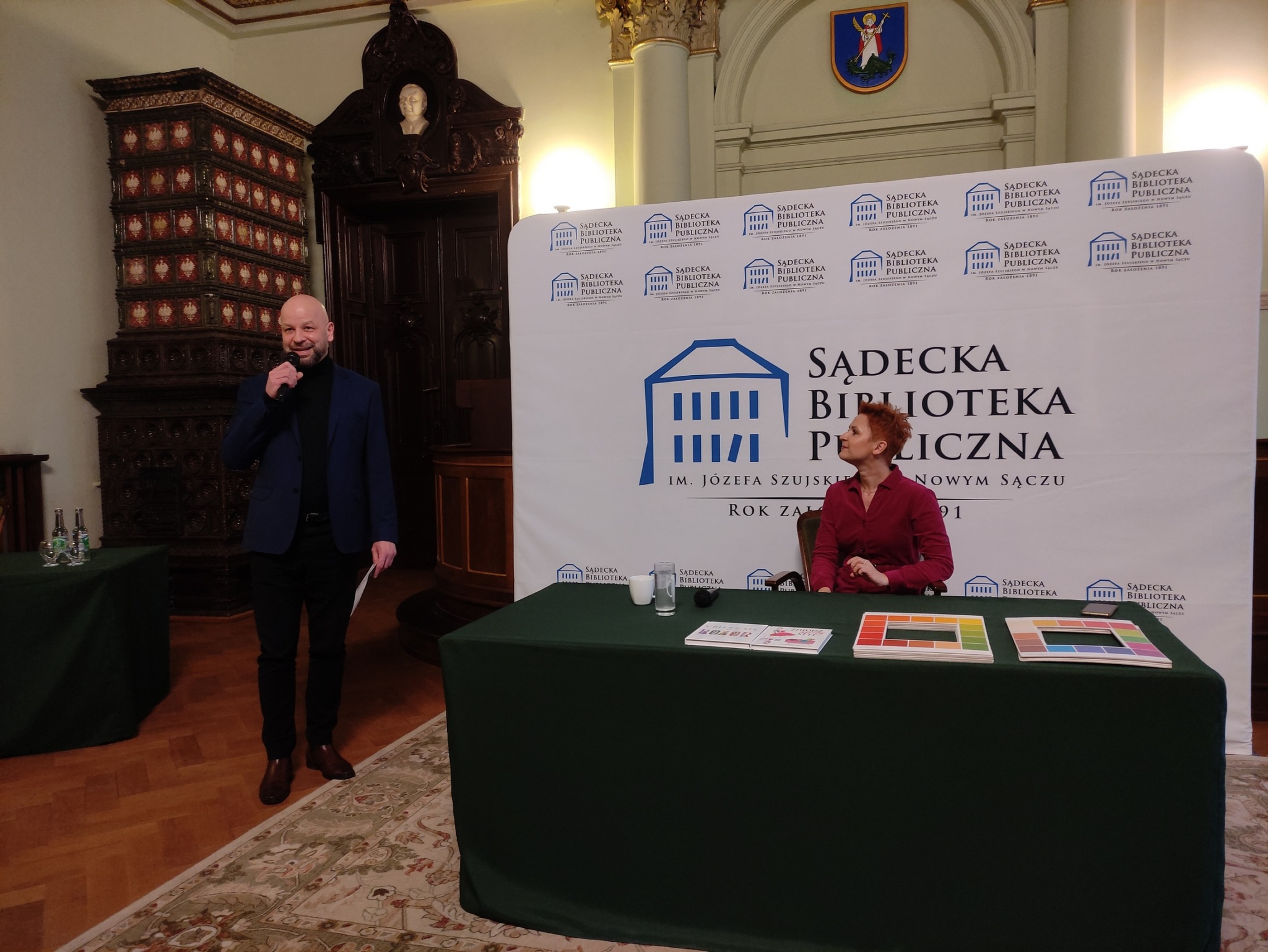Aleksandra Frątczak-Biś_1 Przy dużym, nakrytym ciemnozielonym obrusie stole siedzi kobieta. Spogląda na mężczyznę w granatowej marynarce. Mężczyzna stoi po lewej stronie, przemawia, trzyma w ręku mikrofon.