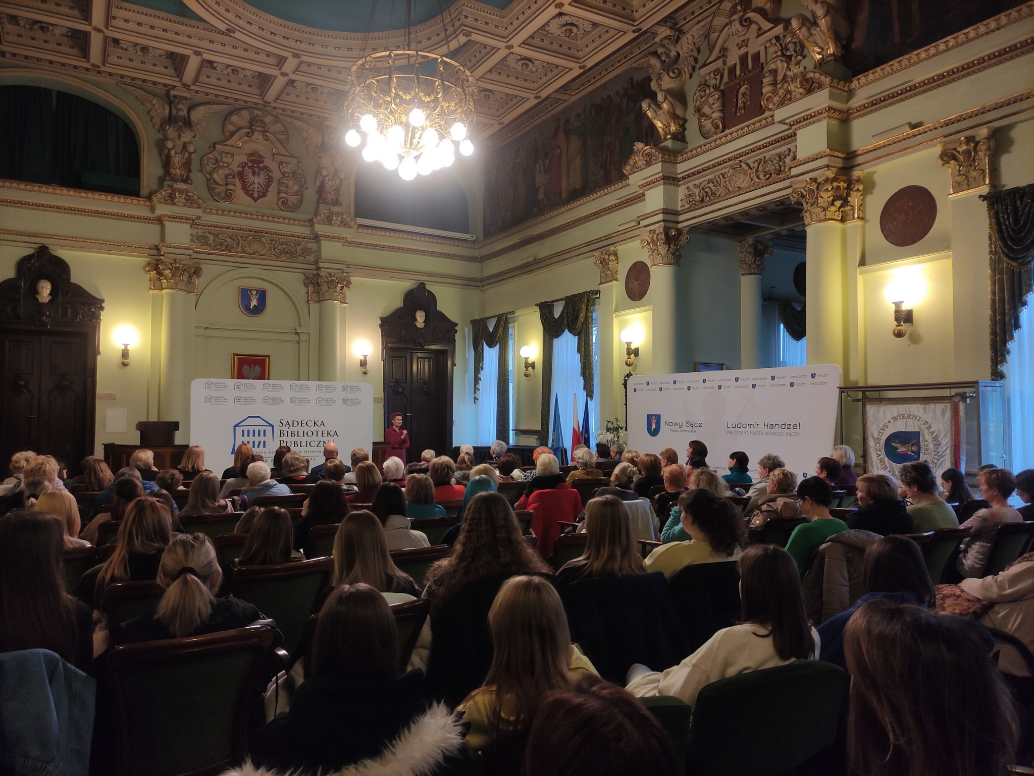 Aleksandra Frątczak-Biś_11 Sala wypełniona jest siedzącymi na widowni ludźmi. W tle stoi przemawiająca do nich kobieta z mikrofonem.