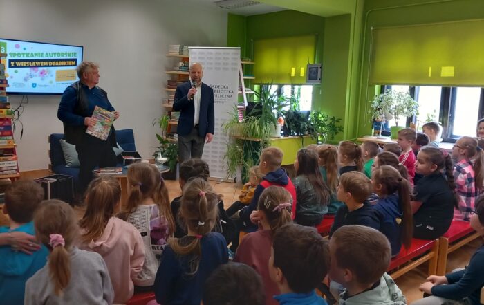 W głębi stoi dwóch mężczyzn, jeden z nich trzyma mikrofon. Za nimi, napis "spotkanie autorskie z Wiesławem Drabikiem" oraz rollup z logotypem biblioteki. Przed nimi na ławkach siedzą zasłuchane dzieci.