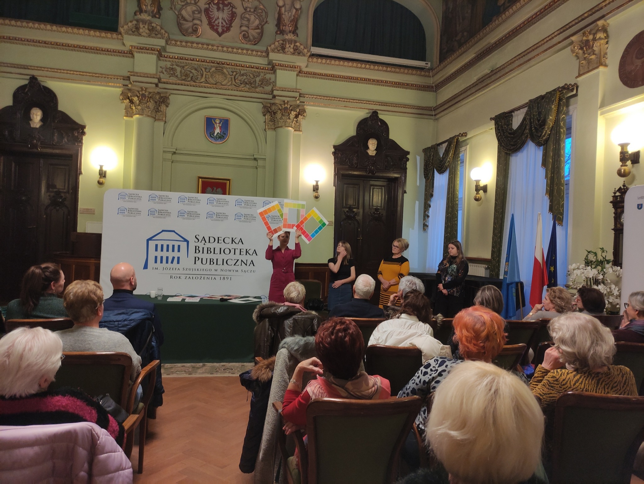 Aleksandra Frątczak-Biś_7 Na widowni siedzą ludzie. Patrzą na cztery zwrócone w ich stronę kobiety. Jedna z nich trzyma wysoko w górze trzy palety z kolorami.