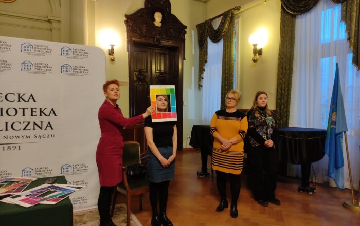 Cztery stojące, zwrócone twarzą do oglądającego kobiety. Jedna trzyma w ręku paletę z kolorami, którą przykłada do twarzy drugiej z nich. Po lewej stronie stół nakryty zielonym obrusem. Leżą na nim palety z kolorami.