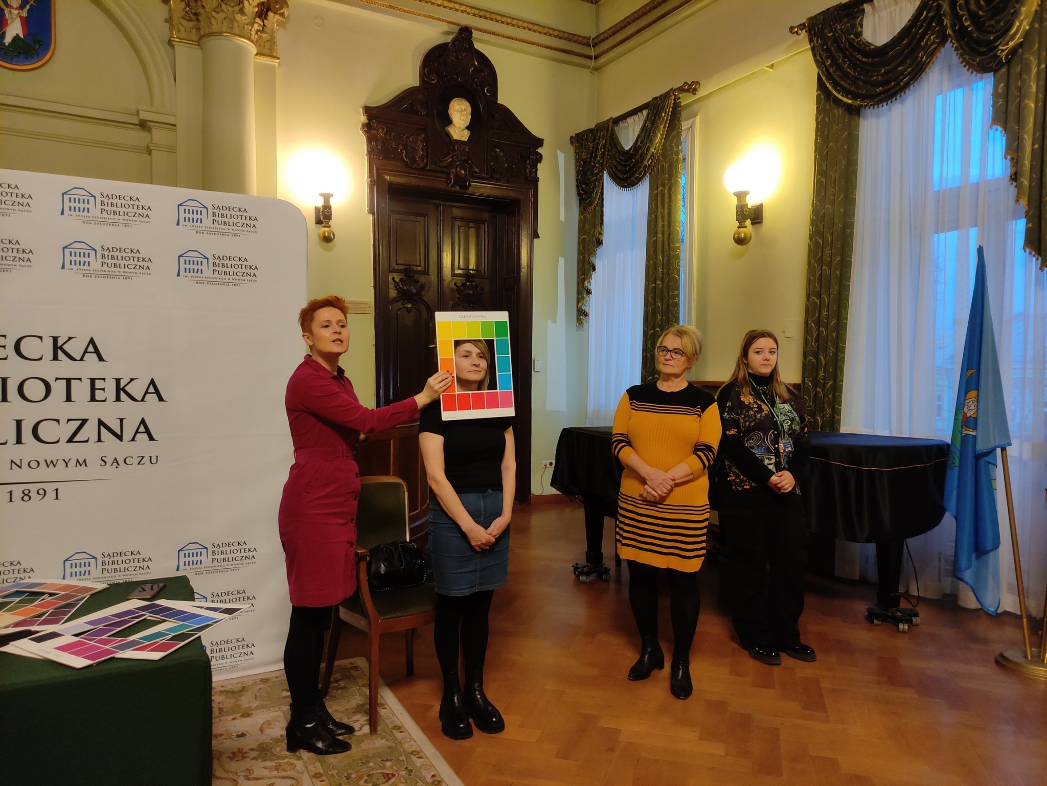 Cztery stojące, zwrócone twarzą do oglądającego kobiety. Jedna trzyma w ręku paletę z kolorami, którą przykłada do twarzy drugiej z nich. Po lewej stronie stół nakryty zielonym obrusem. Leżą na nim palety z kolorami.