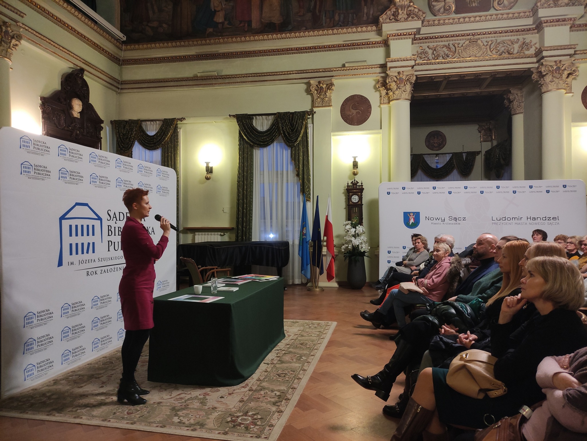 Aleksandra Frątczak-Biś_9 Po lewej stronie obok nakrytego zielonym obrusem stołu stoi kobieta z mikrofonem w ręce. Zwrócona jest w stronę widowni, która zajmuje prawą część zdjęcia.