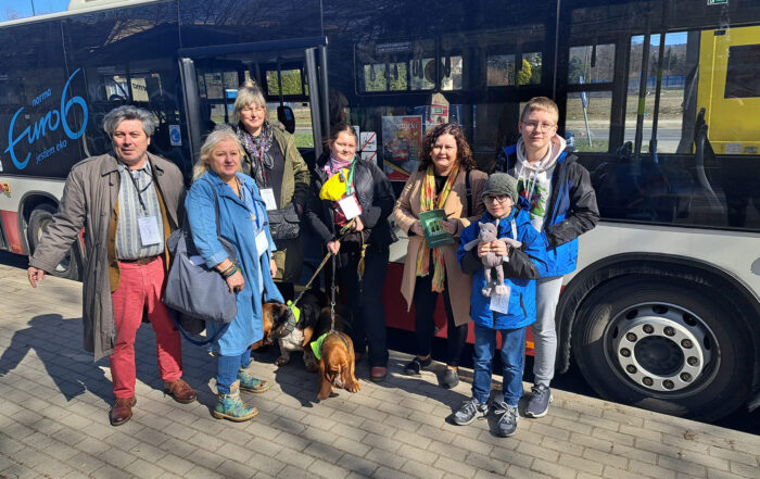 Na chodniku stoi grupa ludzi i dwa psy. Jeden dorosły mężczyzna, trzy kobiety i troje dzieci. Za nimi autobus MPK.