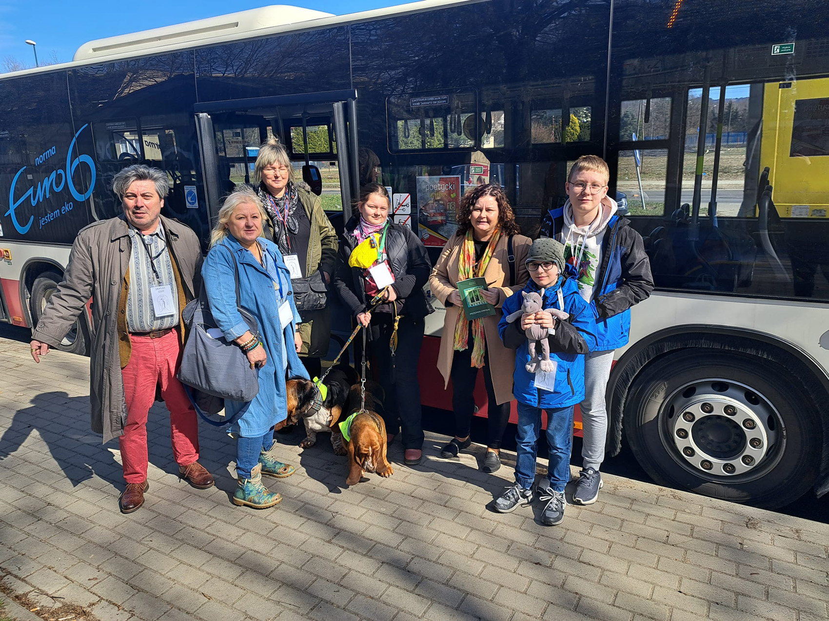 Na chodniku stoi grupa ludzi i dwa psy. Jeden dorosły mężczyzna, trzy kobiety i troje dzieci. Za nimi autobus MPK.