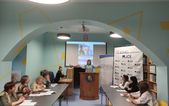 W pomieszczeniu znajduje się 10 osób. Na środku sali stoją naprzeciw siebie dwa długie stoły. Za stołem po lewej stronie siedzą cztery dojrzałe kobiety. Za stołem po prawej stronie siedzi młodzież: trzy dziewczyny i jeden chłopak. Na środku stoi mównica a za nią stoi młoda dziewczyna. Dziewczyna obie ręce ma uniesione, gestykulując. Za mównicą po lewej stronie stoi biurko, a za nim siedzi kobieta. Obie ręce ma złożone i na nich opiera swoją twarz.