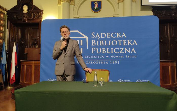 Na zdjęciu stojący mężczyzna. Przed nim stoi duży zielony stół, a na nim butelka wody, szklanka i telefon. Za stołem stoi krzesło. Mężczyzna w prawej ręce trzyma mikrofon, drugą rękę trzyma na oparciu krzesła. Za mężczyzną duża niebieska ścianka: "Sądecka Biblioteka Publiczna".