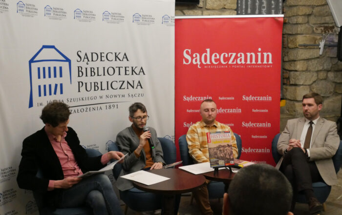 W pomieszczeniu czterech mężczyzn siedzi na fotelach. Przed nimi stoją dwa okrągłe stoliki a na kartki papieru, mikrofon, czasopisma. Mężczyzna pierwszy od lewej trzyma w rękach rozłożone czasopismo, patrzy na nie. Mężczyzna siedzący obok niego trzyma w rękach mikrofon zbliżony do ust. W tle ścianka z napisem "Sądecka Biblioteka Publiczna" a obok niej rollup z napisem "Sądeczanin".