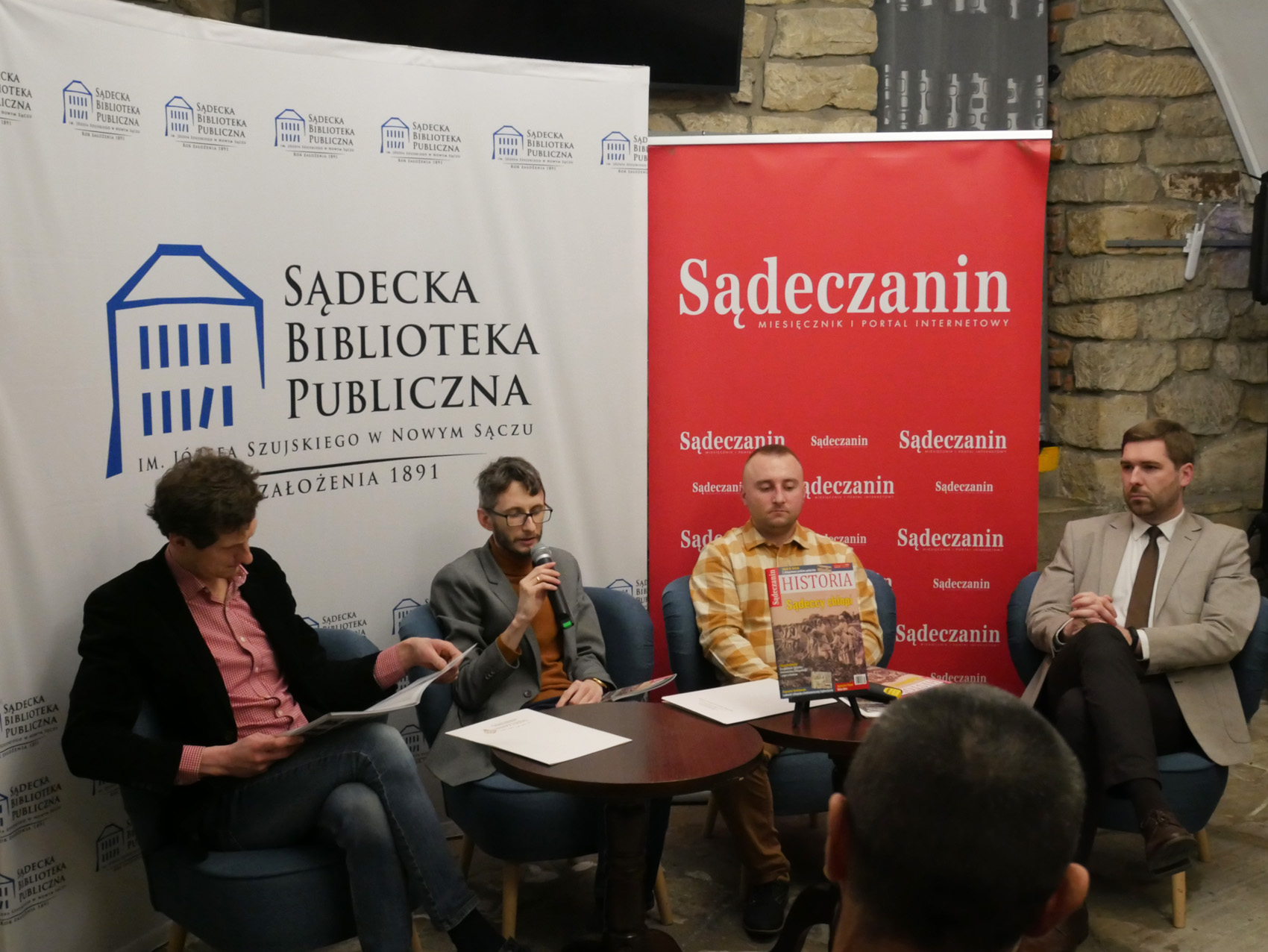 W pomieszczeniu czterech mężczyzn siedzi na fotelach. Przed nimi stoją dwa okrągłe stoliki a na kartki papieru, mikrofon, czasopisma. Mężczyzna pierwszy od lewej trzyma w rękach rozłożone czasopismo, patrzy na nie. Mężczyzna siedzący obok niego trzyma w rękach mikrofon zbliżony do ust. W tle ścianka z napisem "Sądecka Biblioteka Publiczna" a obok niej rollup z napisem "Sądeczanin".