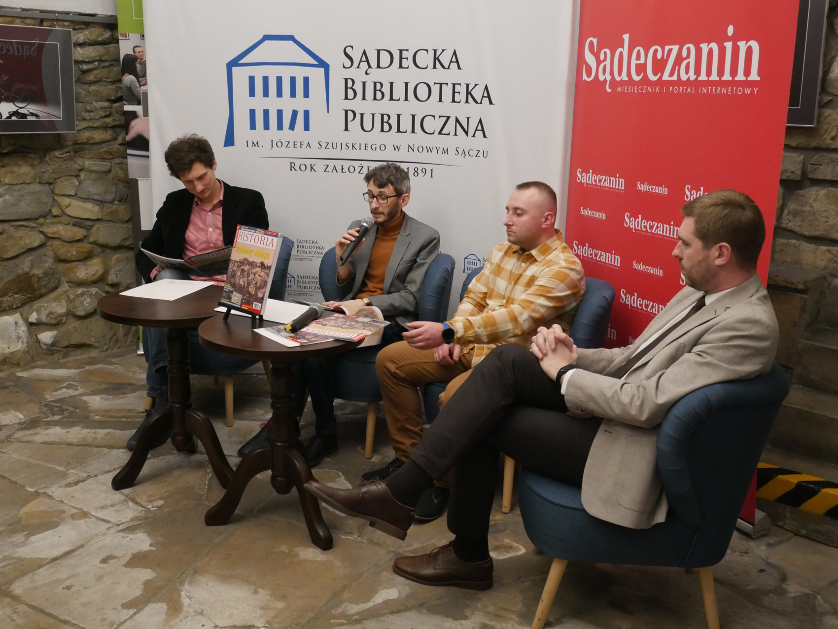 Promocja kwartalnika Sądeczanin Historia (3) W pomieszczeniu czterech mężczyzn siedzi na fotelach. Przed nimi stoją dwa okrągłe stoliki a na kartki papieru, mikrofon, czasopisma. Mężczyzna pierwszy od lewej trzyma w rękach rozłożone czasopismo, patrzy na nie. Mężczyzna siedzący obok niego trzyma w rękach mikrofon zbliżony do ust. W tle ścianka z napisem "Sądecka Biblioteka Publiczna" a obok niej rollup z napisem "Sądeczanin".