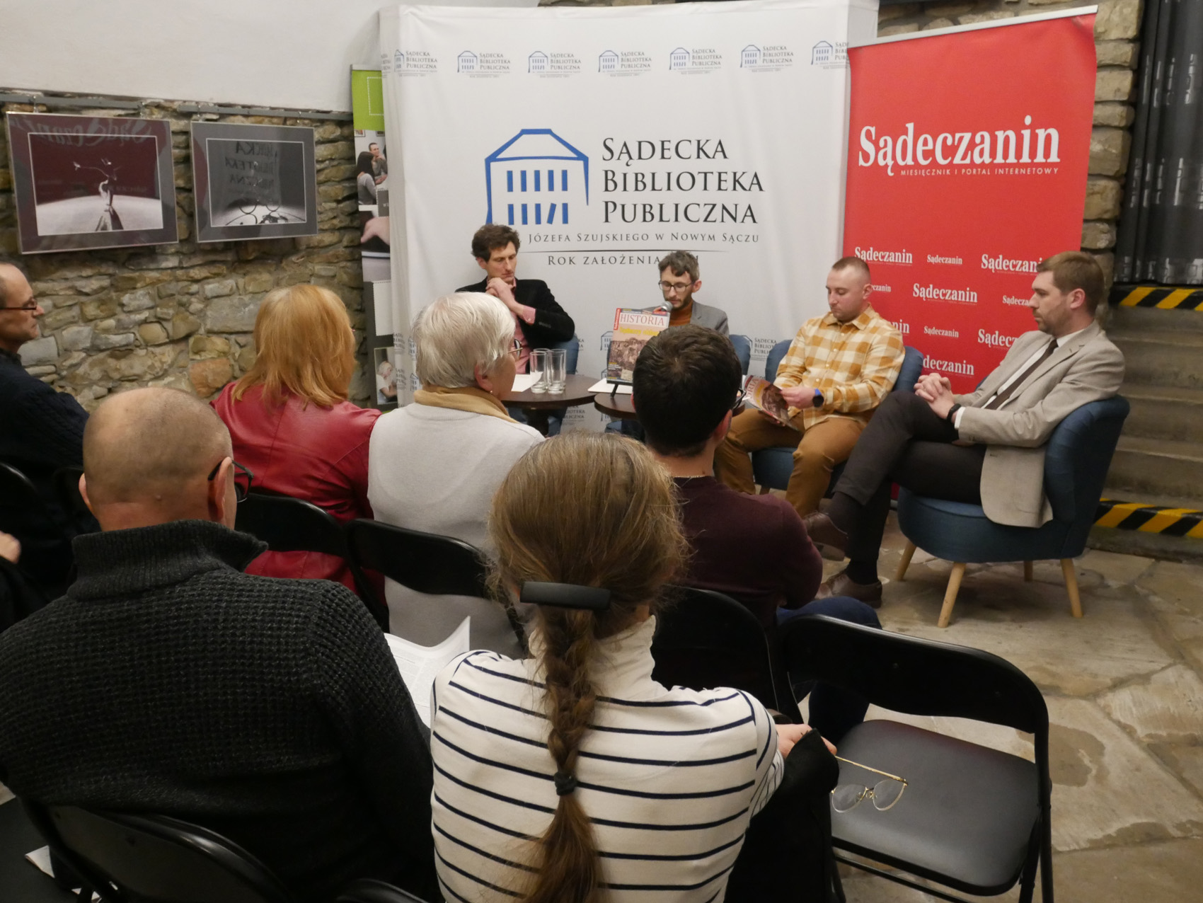 Promocja kwartalnika Sądeczanin Historia (4) W pomieszczeniu grupa siedzących ludzi, pokazana od tyłu. Przed nimi na środku na czterech fotelach siedzi czterech mężczyzn. Za nimi w tle ścianka z napisem "Sądecka Biblioteka Publiczna" a obok niej rollup z napisem "Sądeczanin".