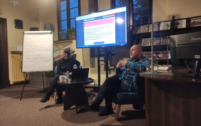 W pomieszczeniu na fotelach siedzą kobieta i mężczyzna. Między nimi stoi mały okrągły stolik a na nim: książka, biały kubek, szklanka, butelka z wodą, laptop. Obok kobiety stoi flipchart z notatkami. Twarz kobiety odwrócona jest w stronę flipchartu, patrzy na niego. Mężczyzna ma obydwie ręce uniesione, gestykuluje. W tle duży ekran, na którym coś jest wyświetlone. Po lewej stronie ekranu stoi duży regał z czasopismami. Po lewej stronie mężczyzny duże brązowe półokrągłe biurko. Na biurku stoją: szklanka, butelka wody, otwarta książka i monitor.