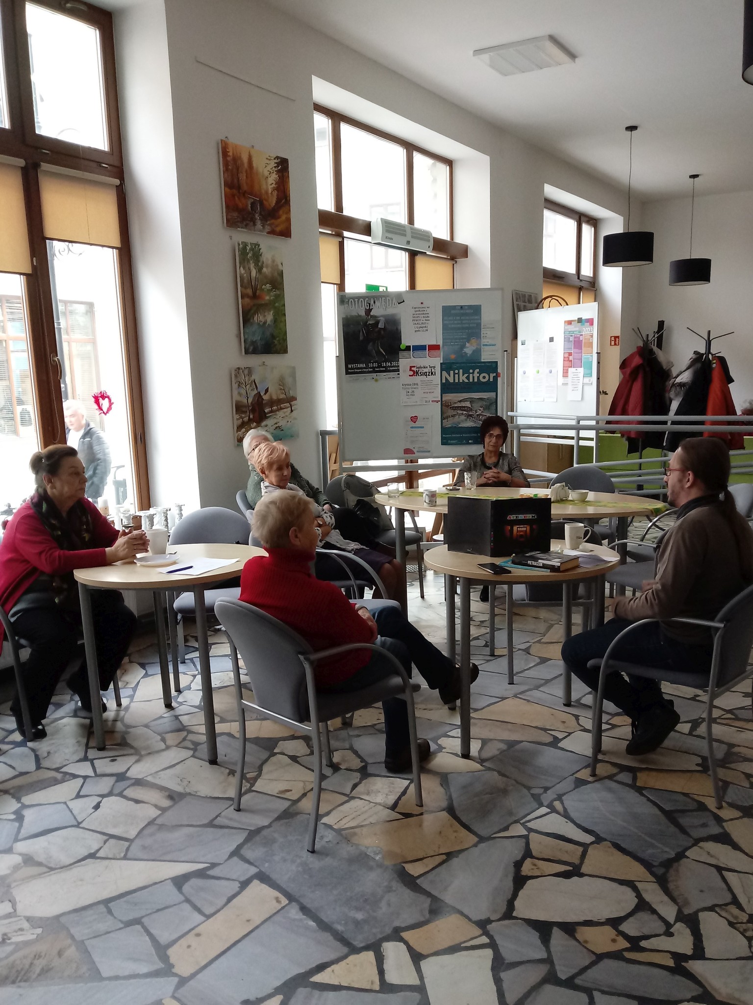 Sroda-z-ksiazka_20230222_1 W pomieszczeniu przy stolikach siedzi sześć osób. Mężczyzna prowadzi spotkanie.