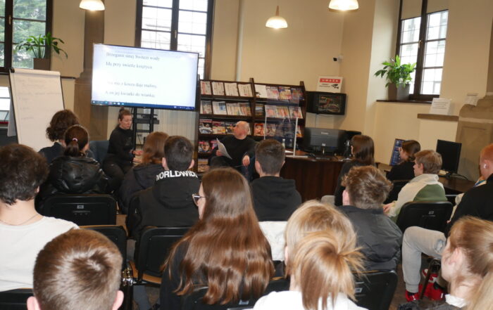 Sala wypełniona ludźmi. Widownia siedzi na krzesłach.Na froncie, zwróceni twarzą do widowni siedzą przy małym stoliku kobieta i mężczyzna. Mężczyzna spogląda na duży telewizor za nimi, na którym wyświetlony jest tekst.