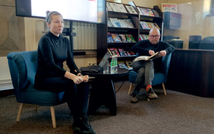 W pomieszczeniu na fotelach siedzą dwie osoby: kobieta i mężczyzna. Po lewej stronie siedzi kobieta i patrzy przed siebie. Obok niej stoi okrągły stolik, na którym stoją: laptop, szklanka, dwie butelki wody. Na fotelu po drugiej stronie stolika siedzi mężczyzna i trzyma w rękach rozłożoną książkę, patrzy na nią.