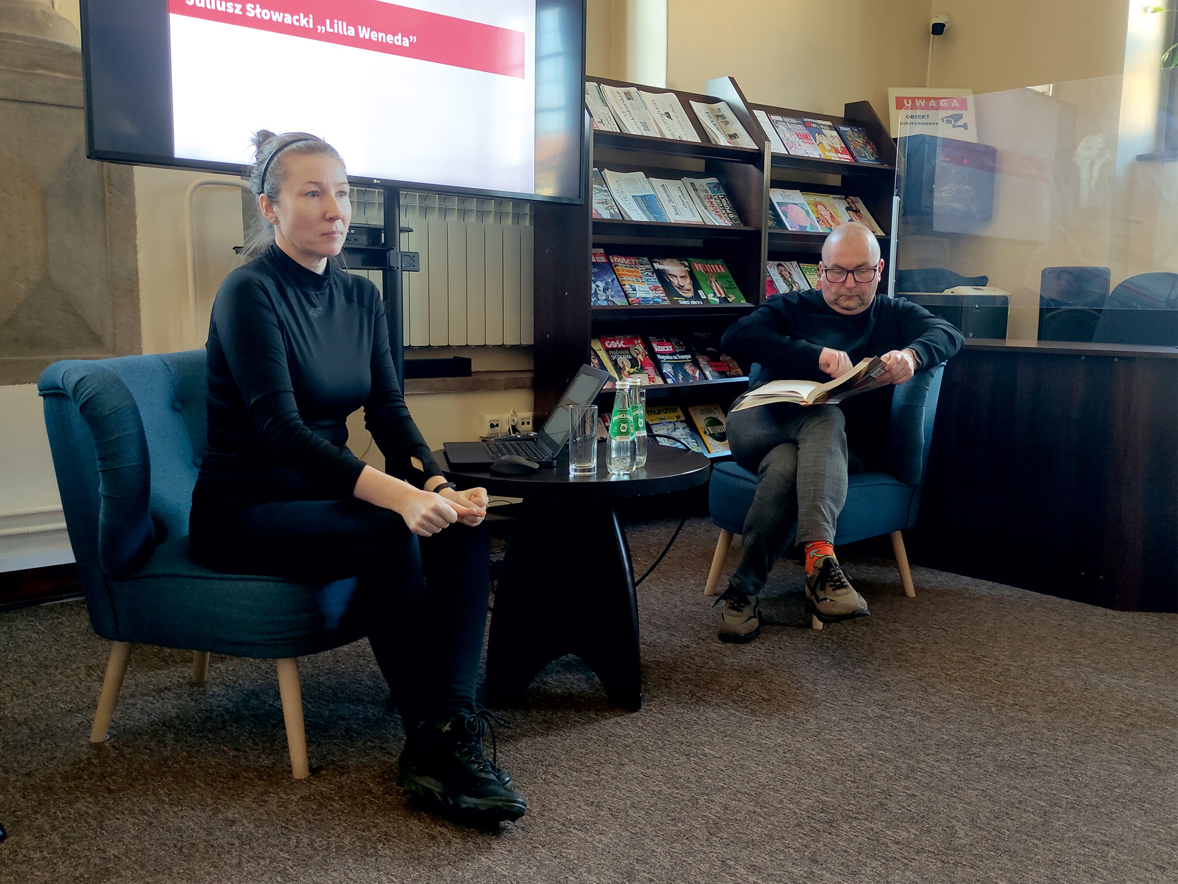 W pomieszczeniu na fotelach siedzą dwie osoby: kobieta i mężczyzna. Po lewej stronie siedzi kobieta i patrzy przed siebie. Obok niej stoi okrągły stolik, na którym stoją: laptop, szklanka, dwie butelki wody. Na fotelu po drugiej stronie stolika siedzi mężczyzna i trzyma w rękach rozłożoną książkę, patrzy na nią.