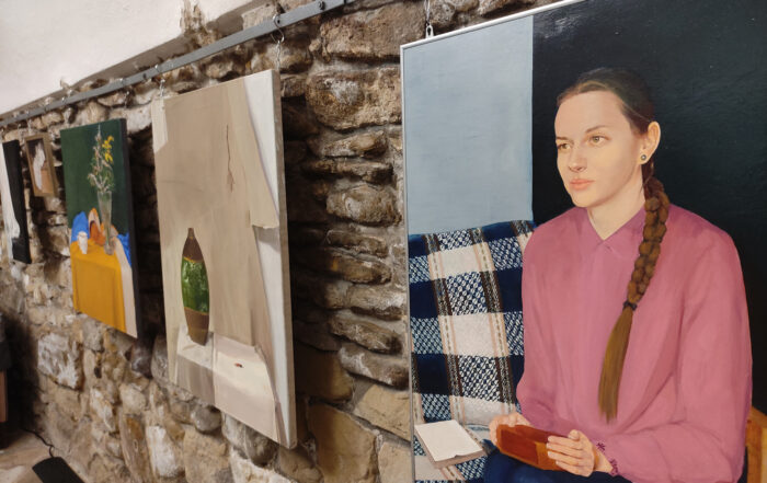 Na murowanej ścianie wiszą kolorowe obrazy. Na pierwszym od lewej obrazie portret młodej dziewczyny z warkoczem. Dziewczyna ubrana jest w różową bluzkę, siedzi na kolorowej kanapie. W rękach trzyma drewnianą szkatułkę, położoną na kolanach. W oddali kolejne obrazy z martwymi naturami.