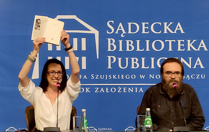 Na zdjęciu kobieta i mężczyzna siedzą na krzesłach. Przed nimi stoi długi stół nakryty zielonym obrusem. Na stole stoją dwie szklanki, dwie butelki z wodą, trzy mikrofony. Kobieta uśmiecha się. Obie ręce ma podniesione do góry, trzyma w nich otwartą książkę.