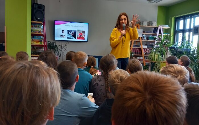 Wnętrze czytelni. Dużo książek i roślin. W tle ekran z napisem "Etapy powstawania książki". Na środku stoi kobieta w żółtym swetrze, mówi do mikrofonu. Przed kobietą siedzą dzieci, słuchają.