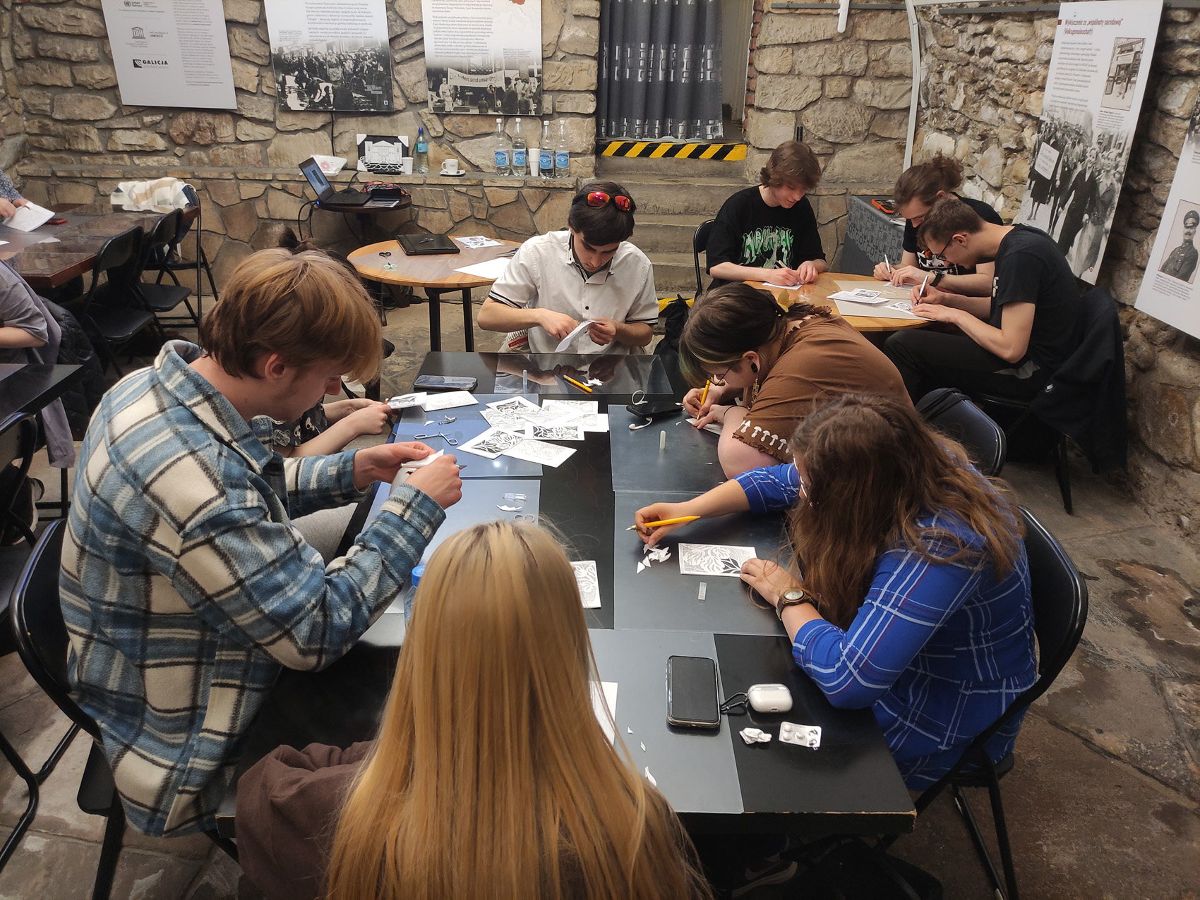 W pomieszczeniu przy stolikach siedzi grupa młodych osób. Na stolikach są rozłożone kartki papieru, nożyki i nożyczki. Siedzące osoby wycinają nożykami wzór na kartce papieru.
