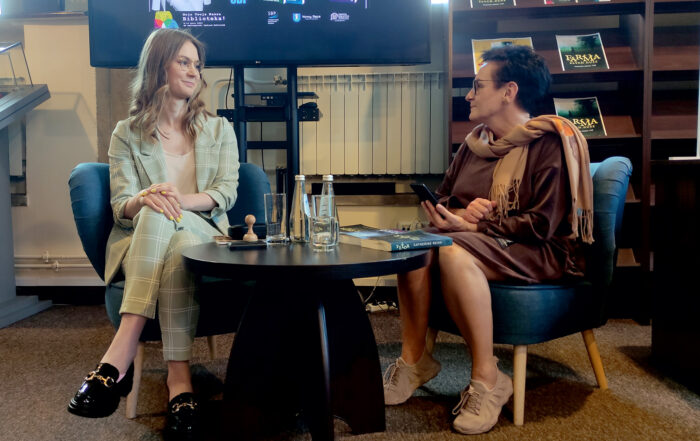 W pomieszczeniu na fotelach siedzą dwie kobiety. Między nimi stoi mały okrągły stolik. Na stoliku stoją: dwie szklanki, dwie książki, dwie butelki wody. Kobiety patrzą na siebie. Kobieta po prawej stronie trzyma w ręce telefon.