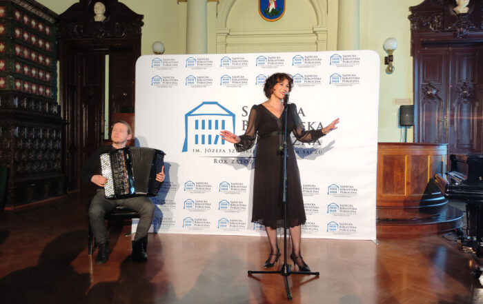 Przy statywie z mikrofonem stoi ubrana w czarną suknię uśmiechnięta kobieta z rozłożonymi rękoma. Obok wpatrzony w dal mężczyzna gra na akordeonie. W tle ścianka reklamowa organizatora z niebieskim domkiem na białym tle.