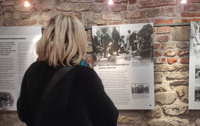 Zwrócona tyłem kobieta z półdługimi blond włosami ogląda wystawiennicze plansze z tekstem i zdjęciami powieszone na ścianie z kamienia.