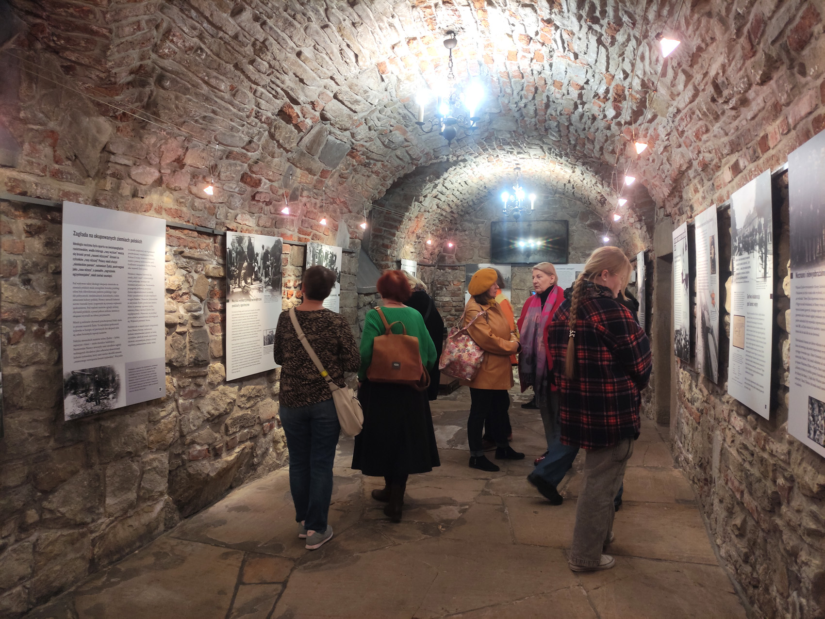 Długa kamienna piwnica ze świecącymi się wiszącymi żyrandolami. W niej grupa ludzi oglądająca plansze wystawiennicze z tekstem i zdjęciami.