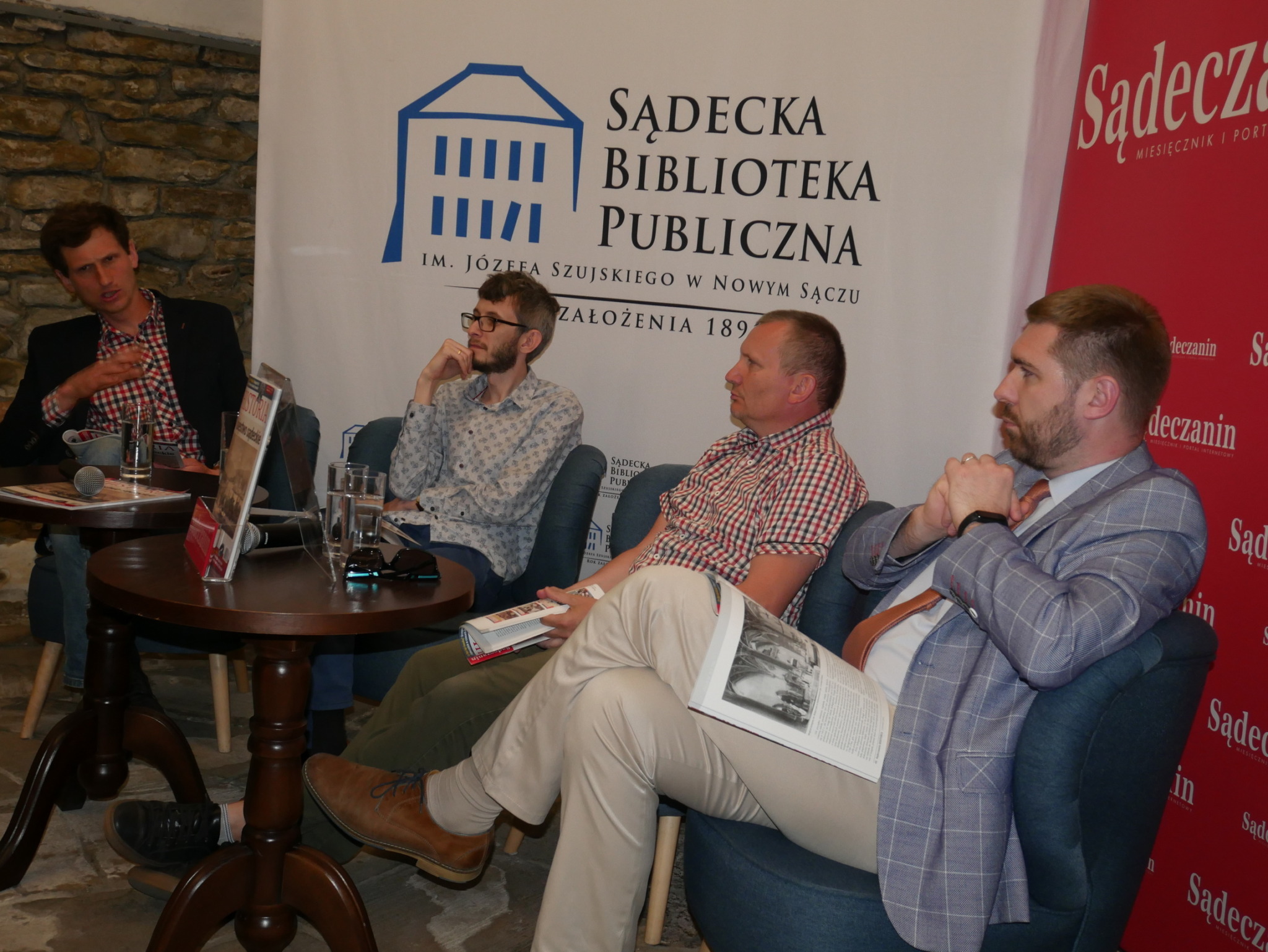 Promocja kwartalnika Sądeczanin HISTORIA_1 Czterech mężczyzn siedzi przy dwóch drewnianych ciemnobrązowych okrągłych stolikach. Za nimi dwie ścianki reklamowe - po lewej stronie biała z niebieskim domkiem i czarnymi napisami a po prawej czerwona z białymi napisami.