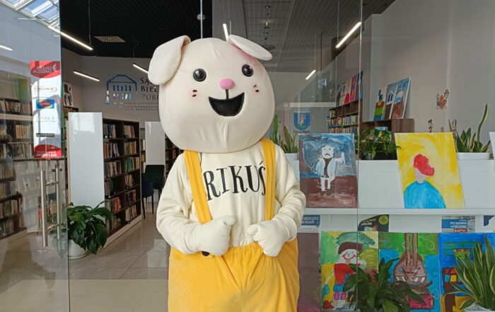 Galeria handlowa. Pośrodku zdjęcia duża "żywa maskotka" - biały króliczek w żółtych spodenkach z napisem na koszulce "Rikuś". W tle przeszklenia i widok na wnętrze nowoczesnej biblioteki.