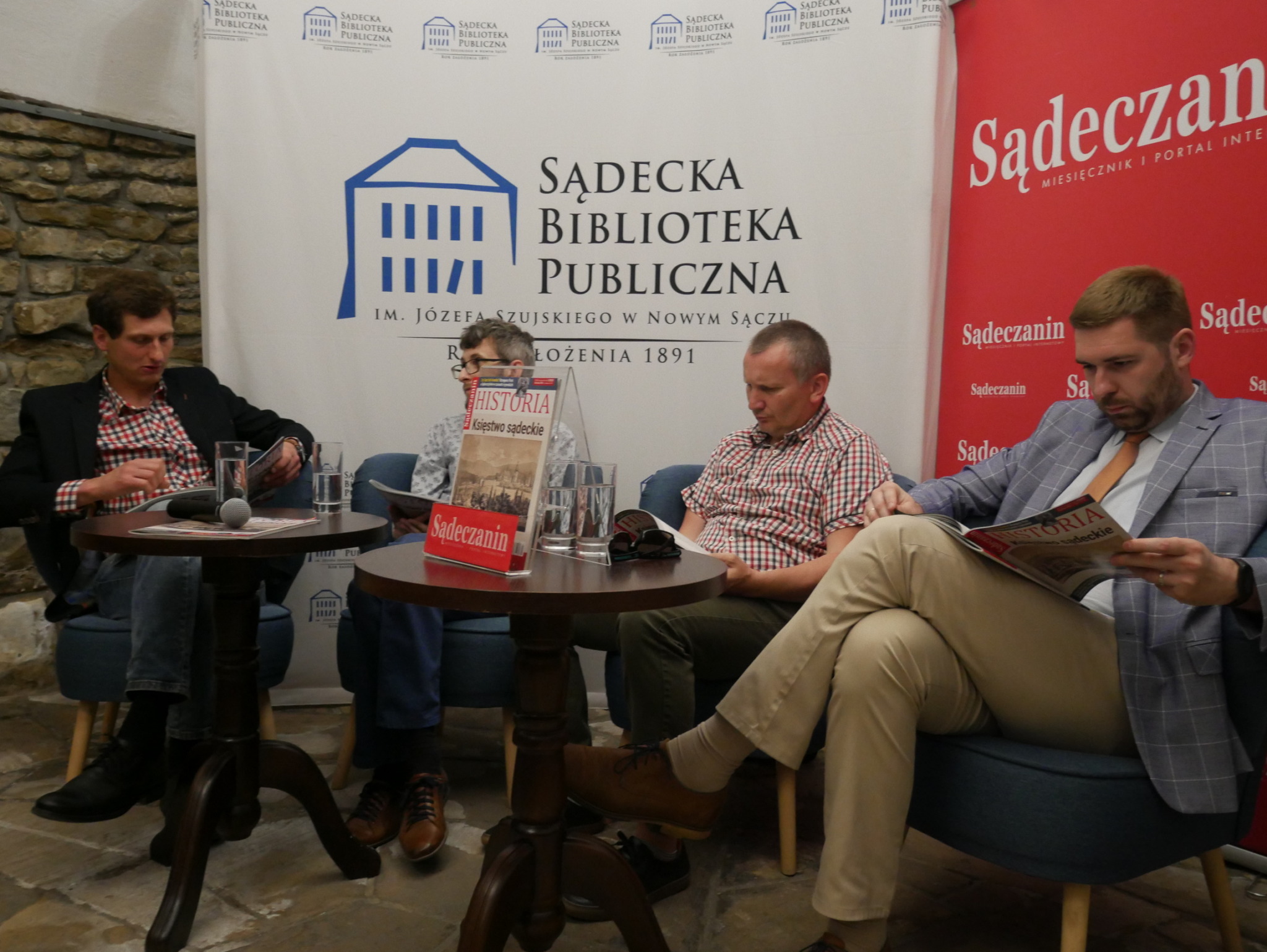 Promocja kwartalnika Sądeczanin HISTORIA_2 Czterech mężczyzn siedzi przy dwóch drewnianych ciemnobrązowych okrągłych stolikach. Czytają kolorową gazetę. Za nimi dwie ścianki reklamowe - po lewej stronie biała z niebieskim domkiem i czarnymi napisami a po prawej czerwona z białymi napisami.