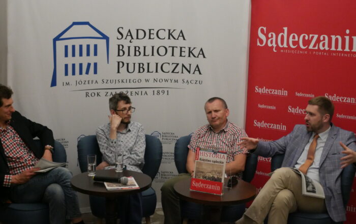 Czterech mężczyzn siedzi przy dwóch drewnianych ciemnobrązowych okrągłych stolikach. Za nimi dwie ścianki reklamowe - po lewej stronie biała z niebieskim domkiem i czarnymi napisami a po prawej czerwona z białymi napisami.