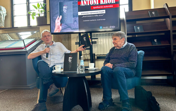 W pomieszczeniu siedzi na fotelach dwóch mężczyzn. Między nimi stoi mały, brązowy, okrągły stolik. Na stoliku znajduje się książka, dwie białe filiżanki, butelka wody. Mężczyzna po lewej stronie w prawej ręce trzyma mikrofon, a w lewej ręce trzyma książkę. Obok siedzącego po prawej stronie mężczyzny na ziemi leży plecak. Za nimi w tle duży ekran.