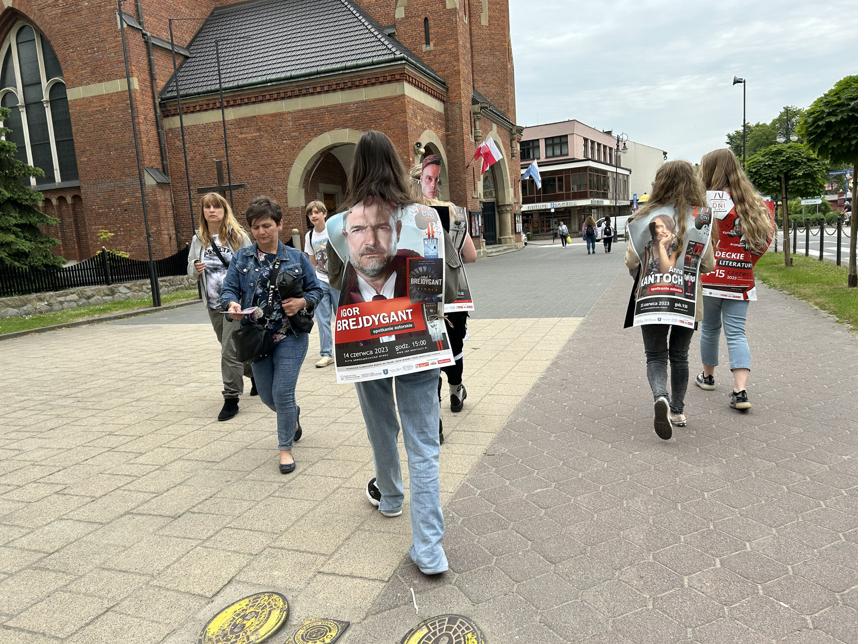 Happening literacki 2023 (11) Osoby ubrane w duże prostokątne reklamy rozdają ulotki przechodniom. W tle po lewej stronie ceglane mury kościoła i ulica a po prawej zielone drzewa.