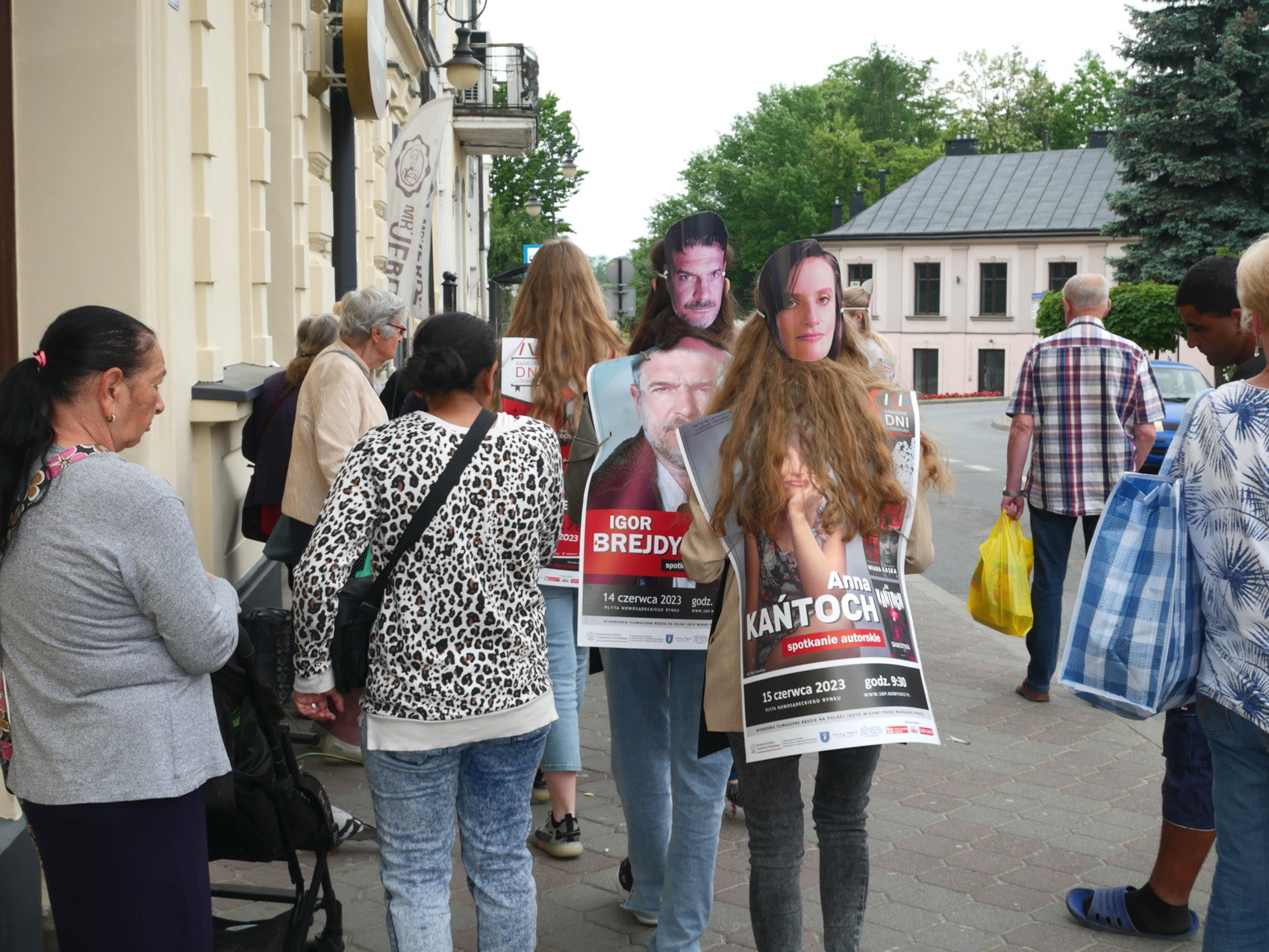 Happening literacki 2023 (3) Grupa ludzi. Wśród nich stojące tyłem, ubrane w duże prostokątne reklamy z maskami z tyłu głowy osoby, które rozdają ulotki.