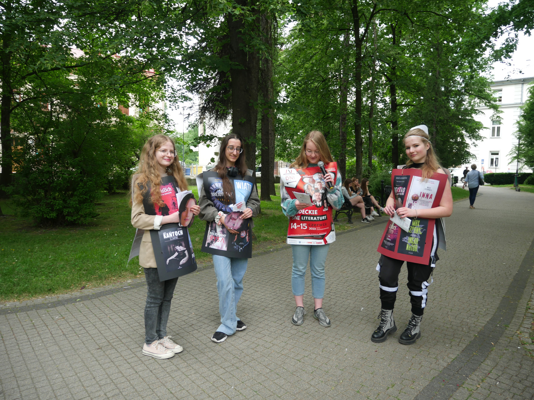 Happening literacki 2023 (4) Cztery młode dziewczyny stoją w parku. Na torsie mają przypięte duże plakaty. W oddali na ławce siedzi grupa młodzieży.