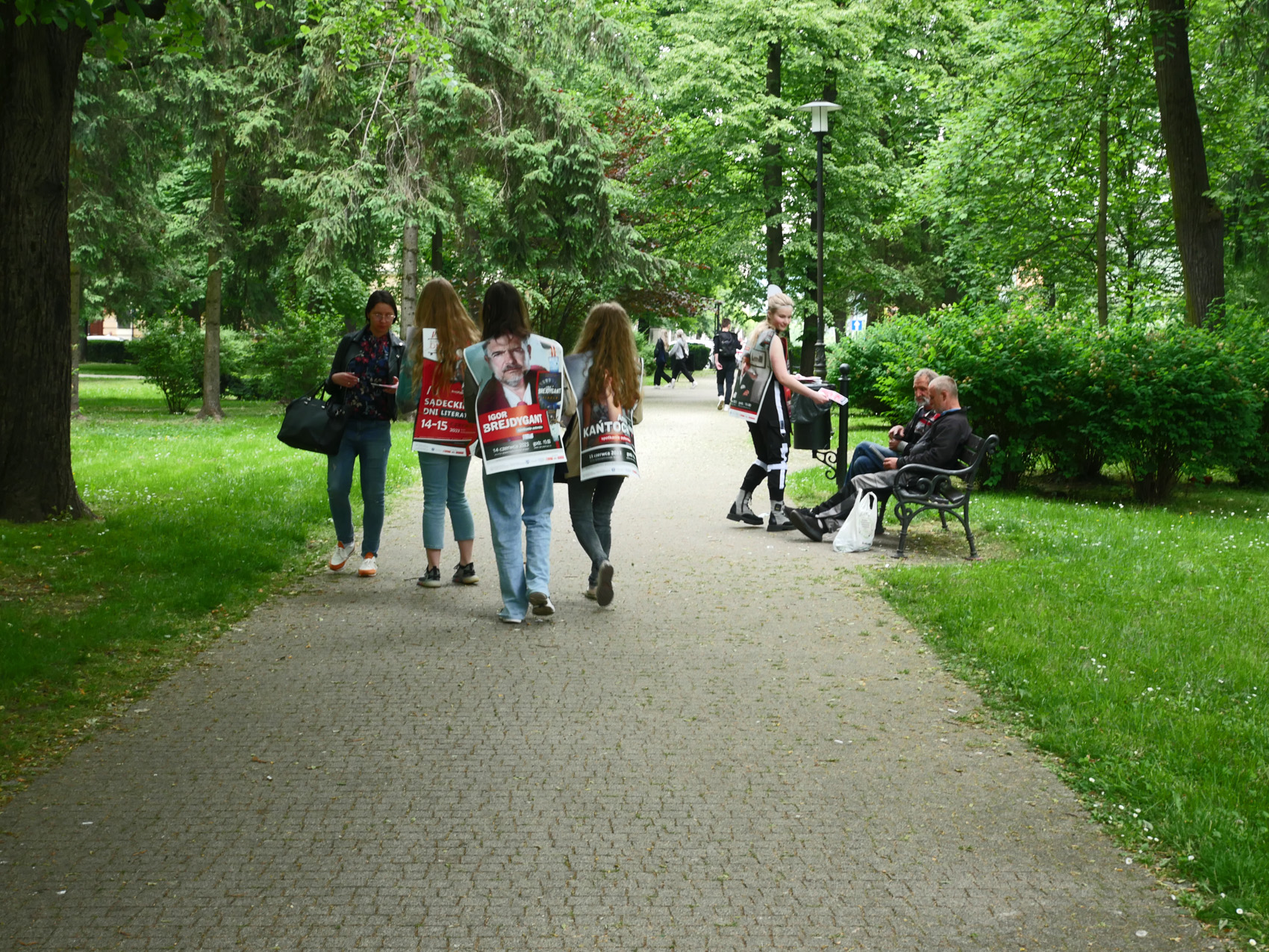 Happening literacki 2023 (5) Grupa osób w parku. Trzy dziewczyny, pokazane od tyłu, mają na plecach zawieszone plakaty. Jedna z nich wręcza ulotkę młodej dziewczynie. W oddali po prawej stronie na ławce siedzi dwóch starszych mężczyzn.