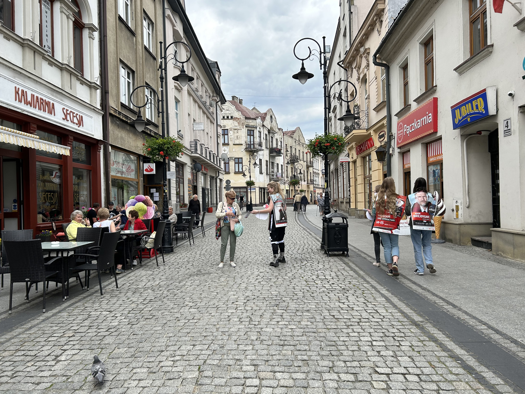 Happening literacki 2023 (7) Brukowana ulica. Po lewej stronie ludzie siedzą w letnim ogródku. W centralnym miejscu osoby z reklamą na plecach rozdają ulotki przechodniom.