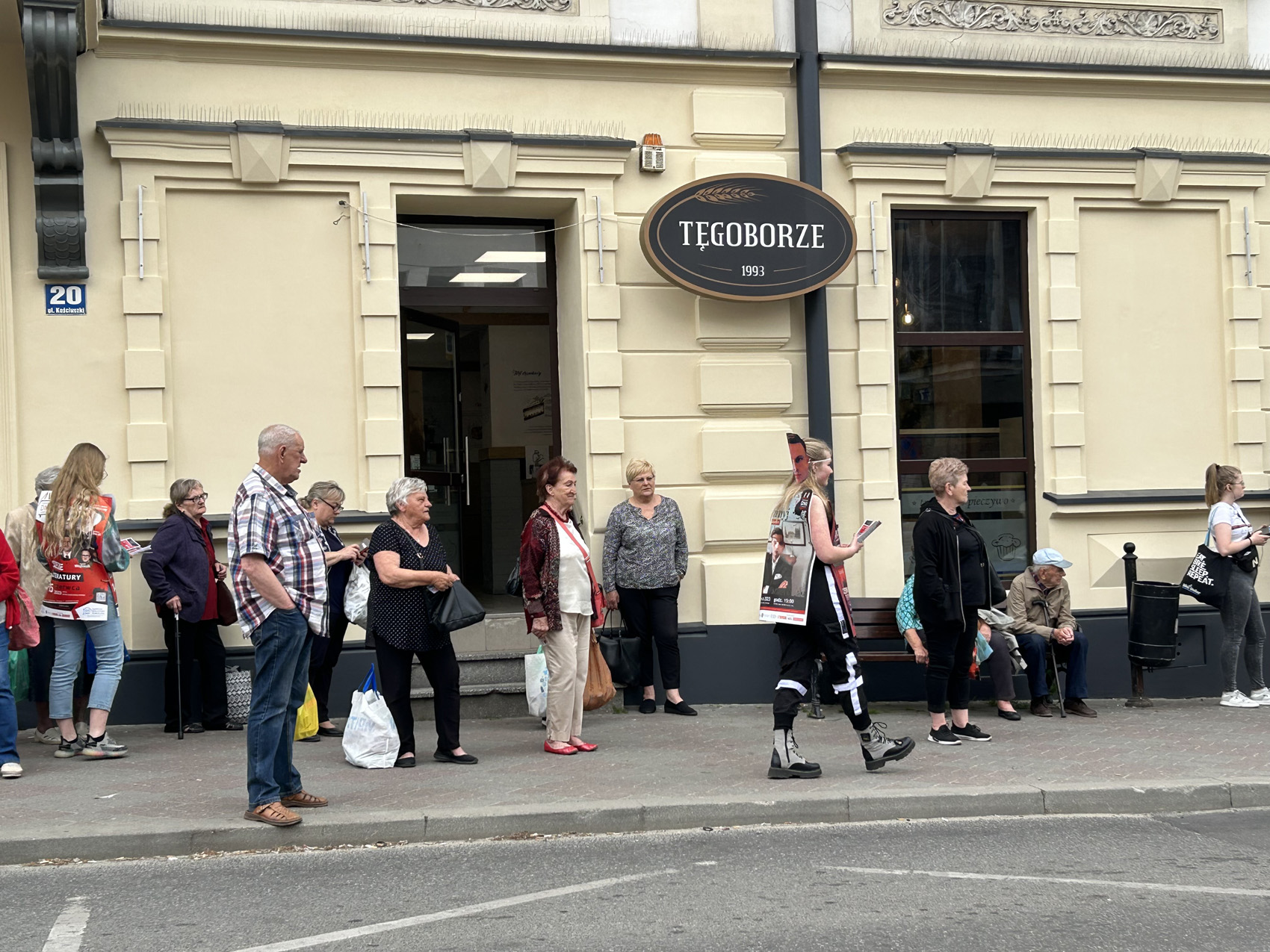 Happening literacki 2023 (8) Kilkanaście osób stoi na chodniku i patrzy na młodą osobę z przypięta z przodu i na plecach reklamą, która trzyma w ręku ulotki. Na pierwszym planie ulica.