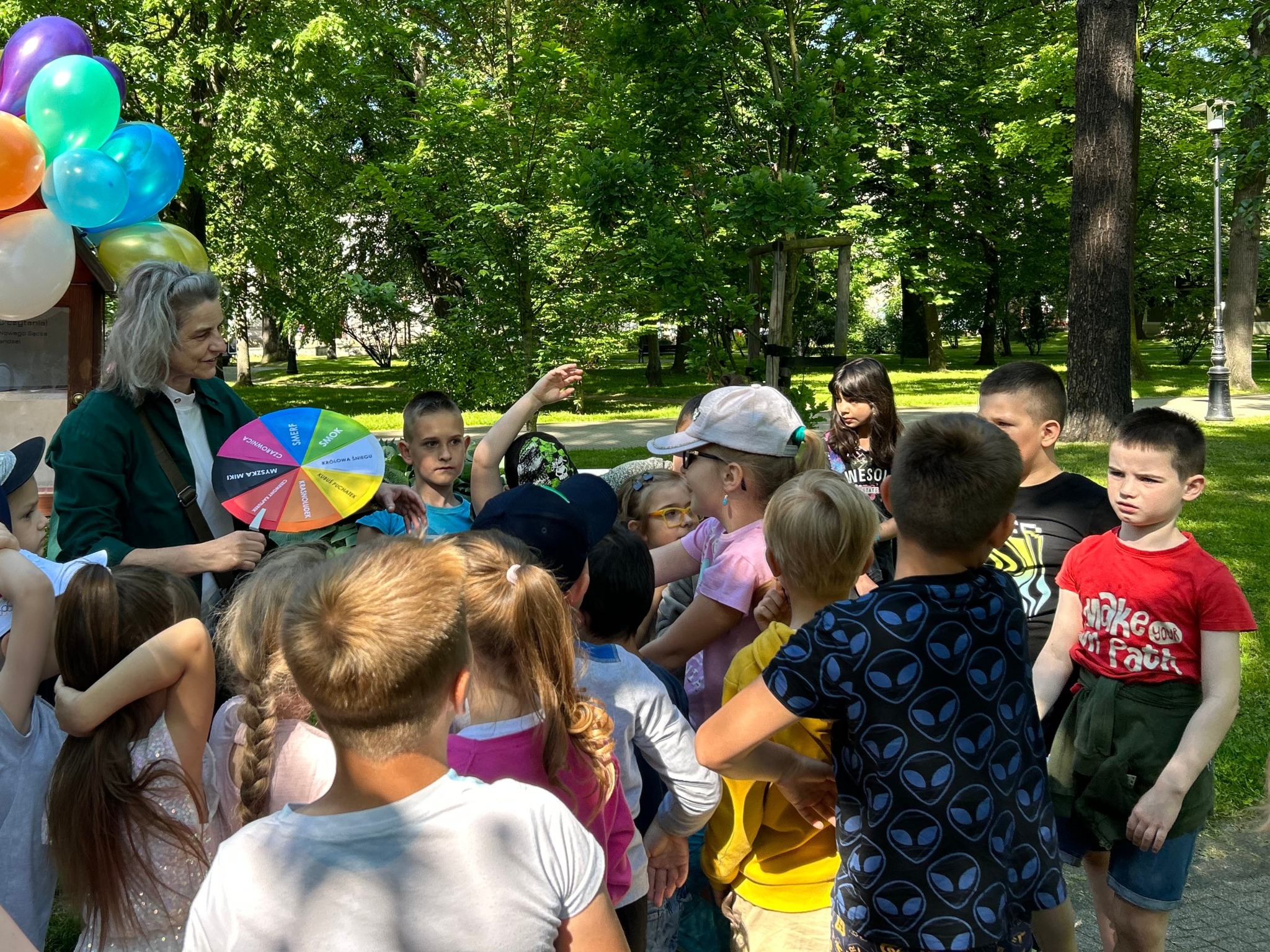 IMG_0099 Park miejski, w tle zielone drzewa liściaste. Kobieta trzyma w rękach kolorowe "koło fortuny". Wokół niej stoi grupa dzieci.